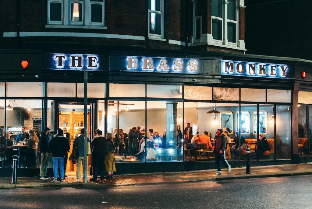 The Brass Monkey on a street corner at night, its illuminated sign lit above windows full of people, with drinkers out on the pavement